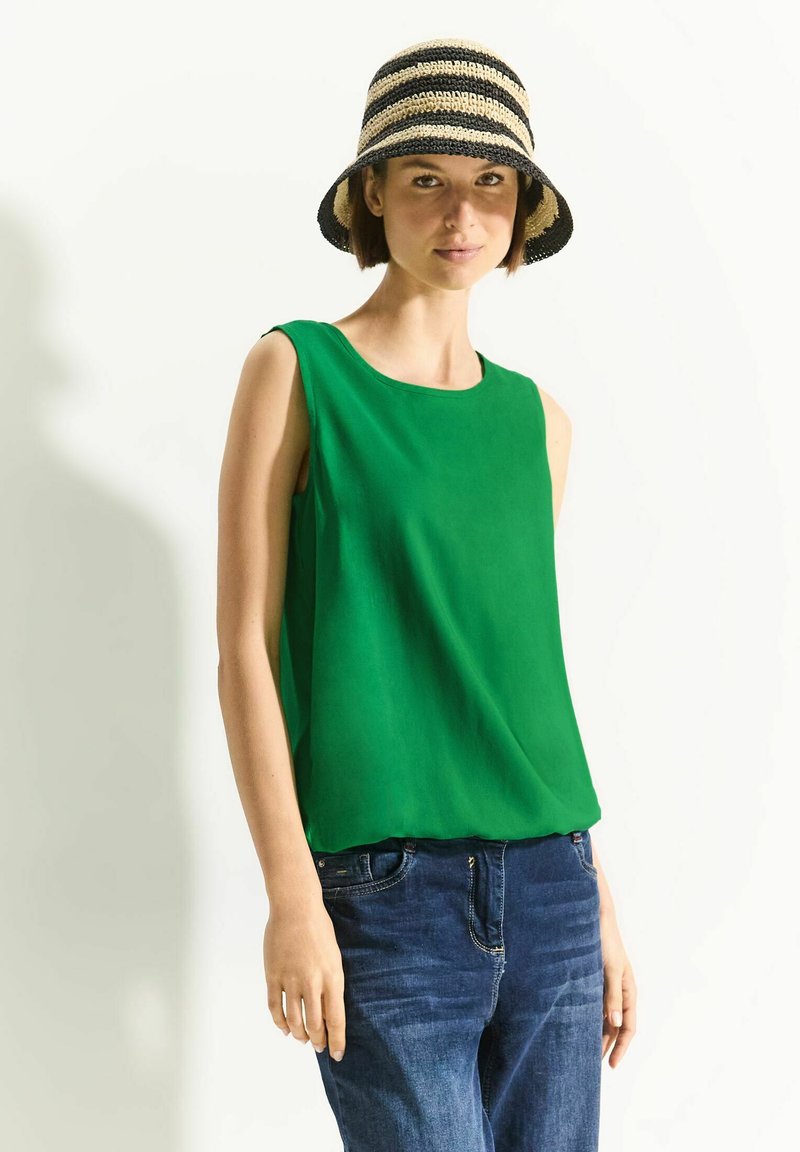 Top sans manches vert en tissu lisse, avec un col rond, associé à un jean en denim bleu foncé et un chapeau de paille à rayures.