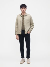 Jeune homme debout portant une surchemise beige boutonnée, un t-shirt vert olive, un pantalon noir retroussé, des chaussettes blanches et des baskets beiges.