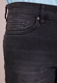 Detailní záběr na černé džínové kalhoty s boční kapsou, na které je malá kovová visačka značky BOSS, a viditelným poutkem na opasek.