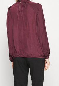 Blusa bordeaux a maniche lunghe con scollatura arricciata, dettaglio a chiave sul retro e orlo elasticizzato, abbinata a pantaloni neri. Tessuto liscio e leggero.