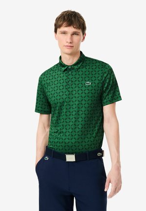 Jonge man die een groen poloshirt met patroon en een donkerblauwe broek met bijpassende riem draagt, staat met één hand in de zak.