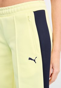 Puma track pants in light yellow with navy side panels. Features a zipper pocket and a small black logo on the left leg. Textured fabric.