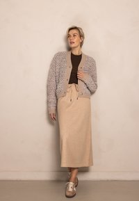 Hellbeiger Cardigan aus strukturiertem Stoff mit geripptem Kragen, kombiniert mit einem langen beigen Rock und braunen Sneakern. Glattes, mehrlagiges Design.