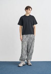 Μαύρο βαμβακερό t-shirt με διακριτικό λογότυπο, συνδυασμένο με άνετο, ασπρόμαυρο παντελόνι με λακκάκια και γκρι αθλητικά παπούτσια. Ουδέτερο φόντο.