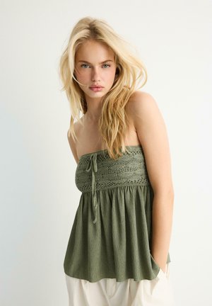 Młoda kobieta o długich blond włosach, ubrana w oliwkowozielony górny top bez ramiączek o fakturze oraz jasne spodnie, stojąca na jednolitym białym tle.