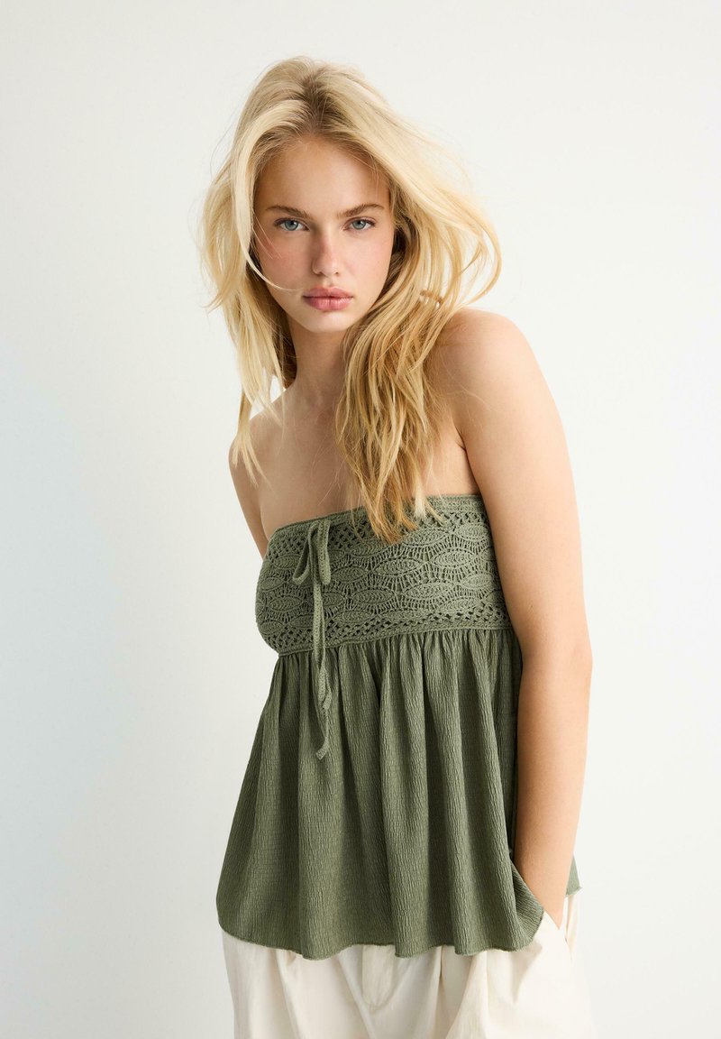 Jeune femme aux longs cheveux blonds portant un haut texturé olive sans bretelles et un pantalon clair, debout devant un fond blanc uni.
