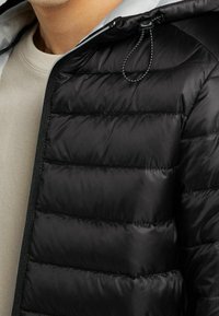 Černá prošívaná bunda s horizontálním prošíváním, šedým vnitřkem a kapucí se stahovací šňůrkou. Má zipové zapínání a hladkou texturu.