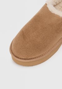 Brown suede slipper with a soft fur lining, rounded toe, and a stitched seam along the sole. Features a flat wooden-like base.