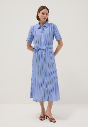 Femme portant une robe midi bleue à rayures verticales, manches courtes, boutonnée, avec une ceinture nouée à la taille et des sandales marron à talons.