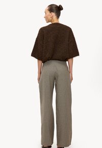 Mujer con el cabello recogido en un moño, lleva pendientes de aro dorados, una blusa marrón de manga corta y textura suave, y pantalones de pierna ancha de talle alto con estampado de cuadros, posando de perfil.