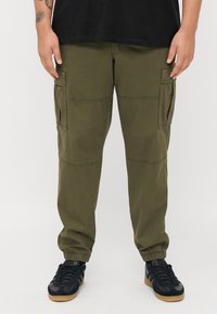 Cargo-pantalons van olijfgroen stof met elastische boorden, meerdere zijzakken en contrasterende stiksels. Gekleed met zwarte sportschoenen.