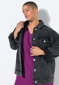 Schwarze Denim-Übergröße-Jacke mit silbernen Knöpfen, Vorderaschen und kontrastierenden schwarzen Manschetten, getragen über einem lila V-Ausschnitt-Kleid.