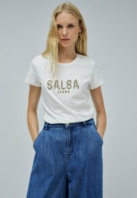 Camiseta blanca con la inscripción "SALSA JEANS" en letras doradas metálicas. Combinada con pantalones de mezclilla azul de tiro alto y corte holgado. Diseño simple y ajuste relajado.