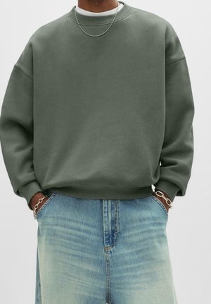 Person iført olivengrøn crewneck sweatshirt, lyseblå jeans, sølvkædehalskæde og armbånd, hænderne stukket i jeanslomme.