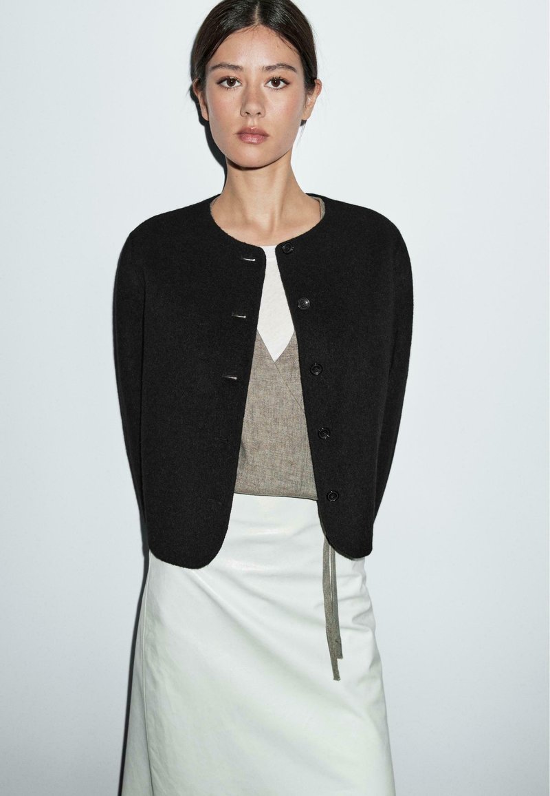 Black, cropped cardigan with a round neckline and five buttons, worn over a beige top and a white leather skirt. Smooth texture.