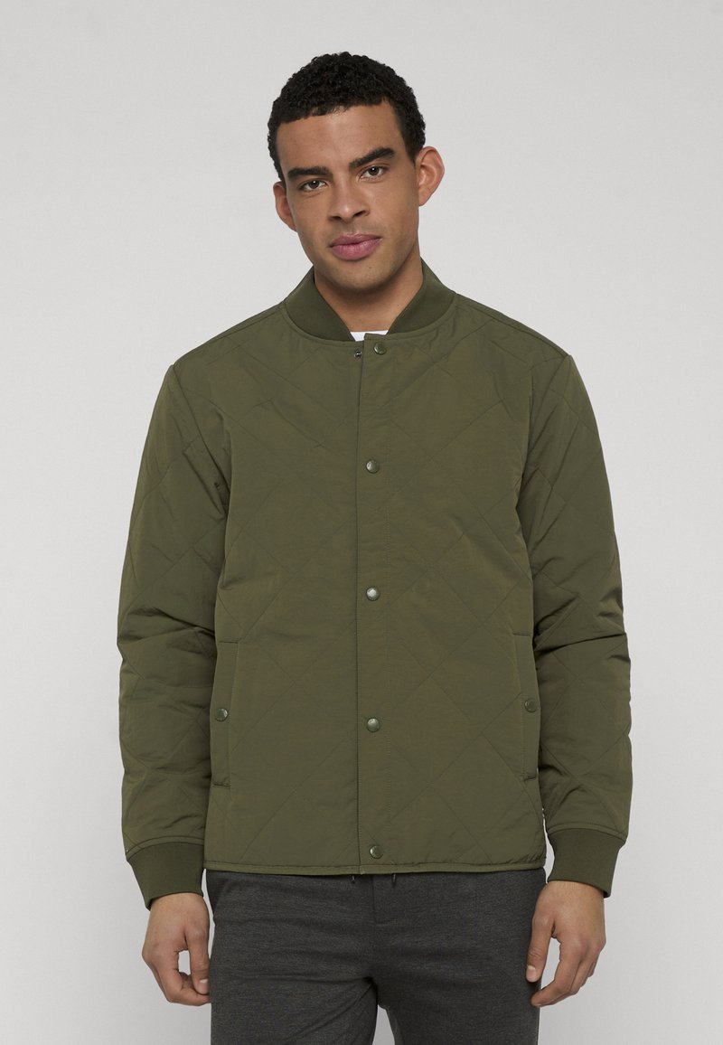 Homme portant une veste matelassée vert olive avec des boutons-pression et des poignets côtelés, debout devant un fond clair uni.