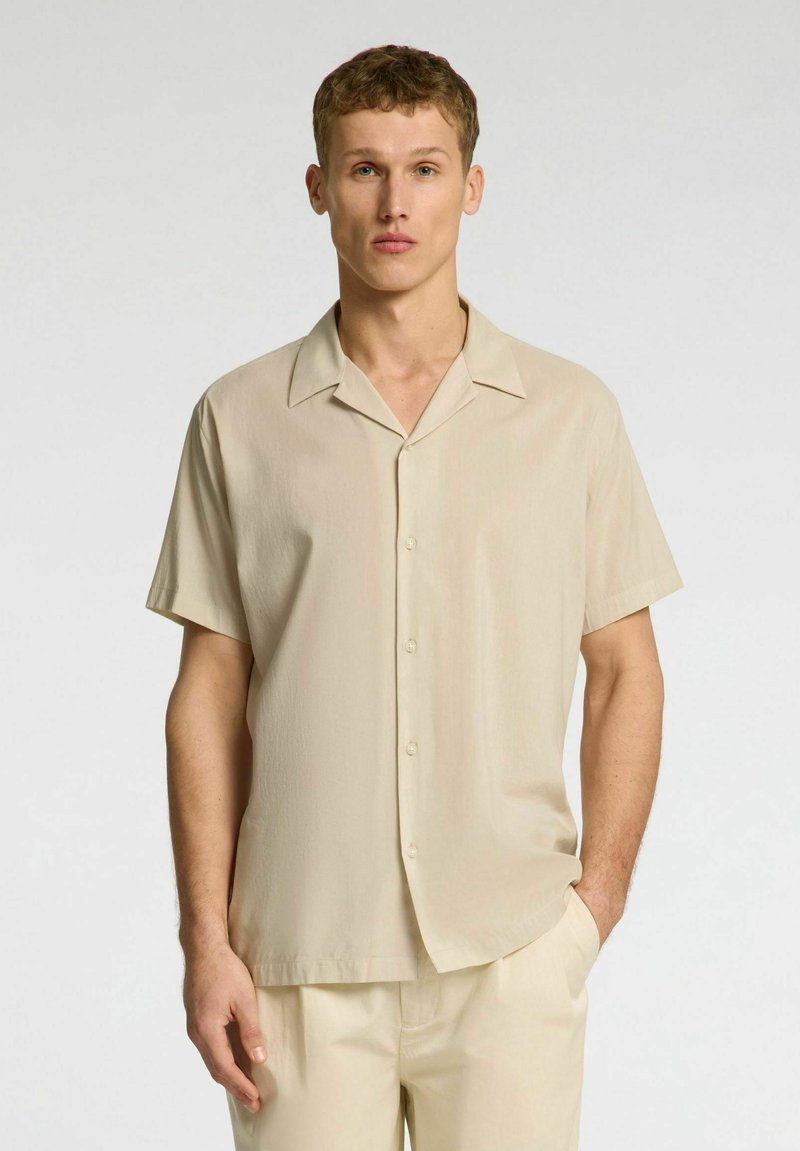 Camicia beige a maniche corte con una texture morbida e liscia. Presenta un colletto, apertura con bottoni e una vestibilità rilassata, abbinata a pantaloni beige coordinati.