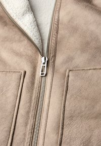 Chaqueta de ante sintético color marrón claro con un zipper plateado, que presenta un suave forro de lana blanca y dos bolsillos delanteros. Superficie texturizada con un acabado liso.