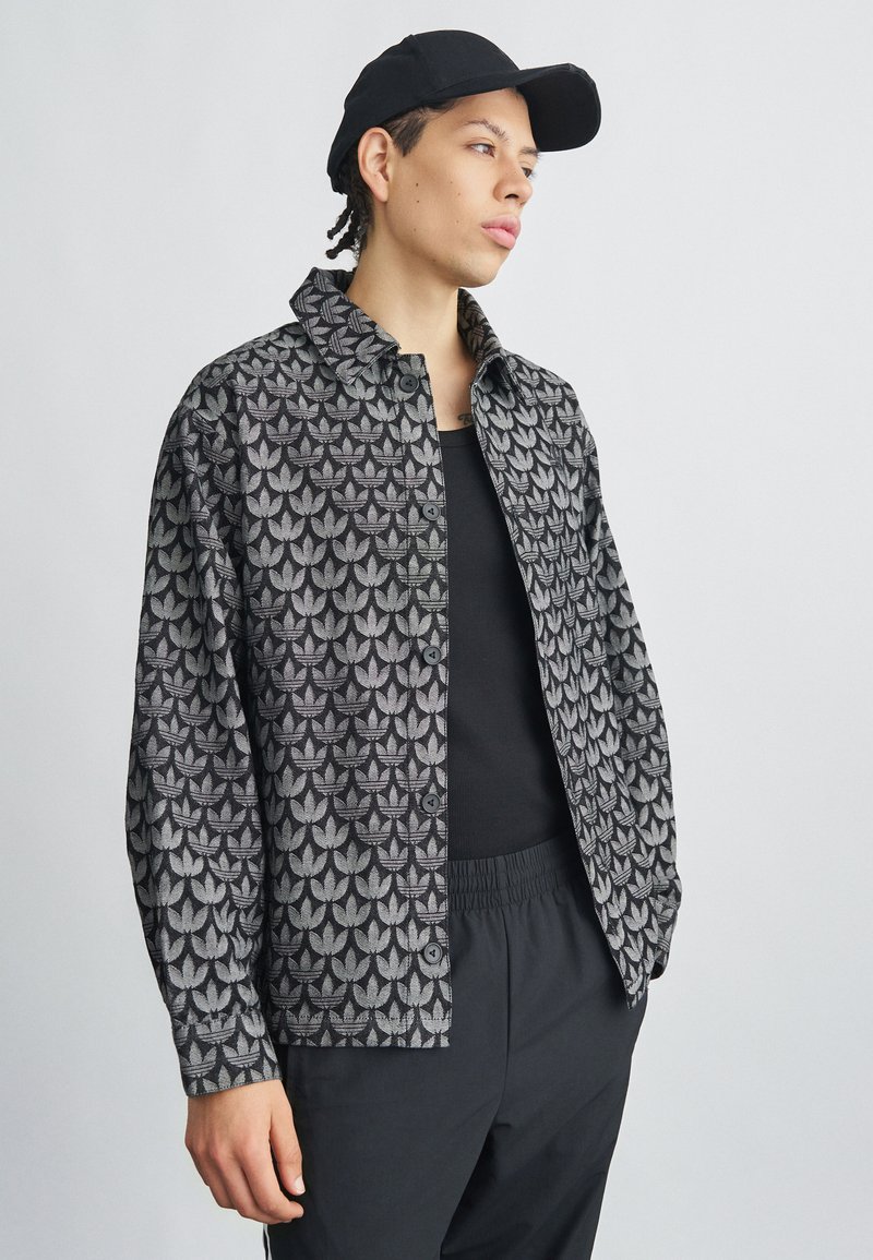 Jaunas vyras, dėvintis juodą kepurę, raštuotą užsegamą striukę, juodą marškinėlį be rankovių ir juodus kelnes, stovintis priešais paprastą šviesų foną.