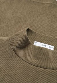 Gros plan d'un pull à col roulé côtelé vert olive montrant la texture et l'étiquette de taille à l'intérieur du col.