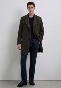 Abrigo de mezcla de lana marrón con un diseño ajustado, solapas anchas y botones de un solo pecho, combinado con pantalones azul marino y zapatos negros.