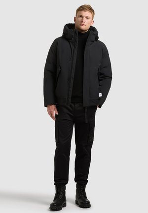 Un homme porte une veste noire à capuche, un pantalon cargo noir et des bottes à lacets sur un fond clair et uni.