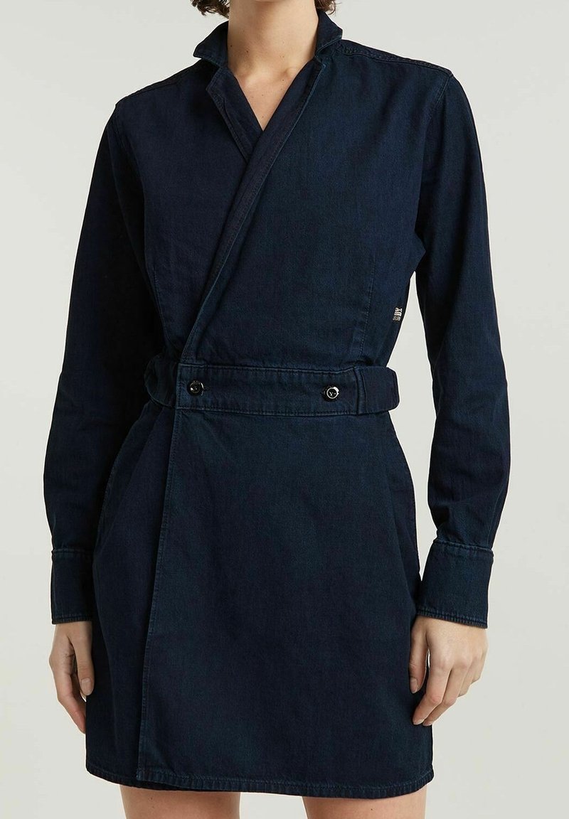 Femme portant une robe en jean bleu foncé avec manches longues, devant croisé et ceinture boutonnée à la taille.