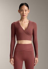 Donna che indossa un top a portafoglio corto a maniche lunghe color ruggine abbinato a leggings a vita alta, in piedi contro uno sfondo neutro.