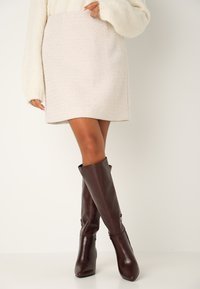 Pletená krémová mini sukně s texturovaným povrchem doplněná o těsné hnědé kožené vysoké boty po kolena s špičatou špicí a mírným leskem.