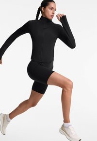 Giacca sportiva nera con colletto alto e maniche lunghe, abbinata a pantaloni corti da ciclismo neri. Sneakers bianche completano il look sportivo.