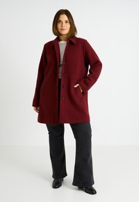 Abrigo de mezcla de lana burdeos con cierre de botones, cuello puntiagudo, bolsillos laterales y forma estructurada, combinado con jeans de campana negros y botas hasta el tobillo.