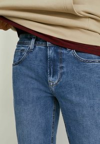 Jean en denim bleu délavé avec une coupe droite, présentant un design à cinq poches et des coutures contrastantes, visibles sous un haut en couches beige et bordeaux.