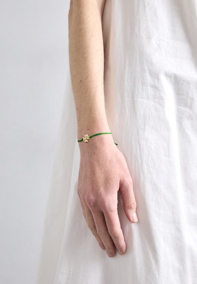 Green string bracelet with a small gold flower charm, worn on a light-skinned, slender wrist against a white background.