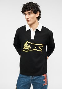 Μαύρο πόλο με μακριά μανίκια και λευκό γιακά, με μεγάλο κίτρινο σχέδιο καρτούν ζώου στο μπροστινό μέρος. Φοριέται με μπλε τζιν παντελόνι.