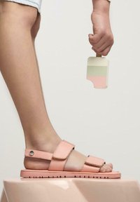 Sandalias rosas con dos correas ajustables, suela plana y suela texturizada. Mano sosteniendo un helado de colores múltiples. Fondo neutro.