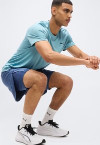 Άνδρας με μπλε αθλητική μπλούζα και σορτς εκτελεί άσκηση καθίσματος φορώντας λευκά αθλητικά παπούτσια Puma και κάλτσες, μπροστά σε μονόχρωμο φόντο.