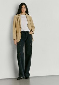 Femme aux longs cheveux noirs portant un blazer beige, une chemise blanche, un jean noir à jambes larges avec des découpes florales, et des chaussures à talons noires, debout contre un mur.