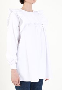 Weißes Langarmbluse mit Rüschenschultern, gerafftem Oberteil und eng anliegenden Manschetten, kombiniert mit dunkelblauen Jeans. Glattes Material mit strukturierter Form.
