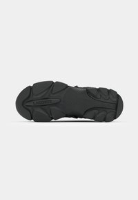 Suela de zapatilla negra con goma texturizada, que presenta un patrón en zigzag y protuberancias redondeadas. Incluye "LACOSTE" en relieve en el centro.