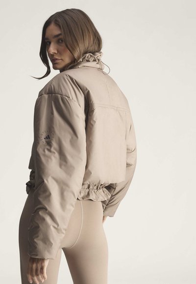 Frau, die sich umdreht, trägt eine beige gekürzte Steppjacke und passende Leggings vor hellem einfarbigem Hintergrund.