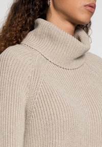 Beżowy sweter dzianinowy z wysokim, złożonym kołnierzem. Teksturowany materiał z prążkowaniem i luźnym krojem, prezentujący zbliżenie na dekolt.