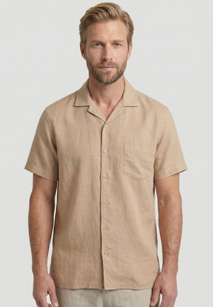 Volwassen man met kort blond haar en baard, die een beige overhemd met korte mouwen en borstzak draagt, tegen een effen achtergrond.