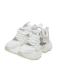 White sneakers with silver accents, featuring a smooth leather upper, thick rubber sole, lace closure, and a branded metal charm.
