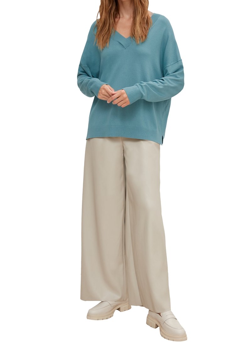 Femme portant un pull en V couleur sarcelle, un pantalon beige à jambes larges et des mocassins crème, debout avec les mains légèrement jointes.