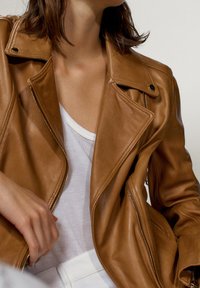 Giubbotto biker in pelle marrone con dettagli in zip argentati, revers a punta e una texture liscia, indossato sopra una canottiera bianca.