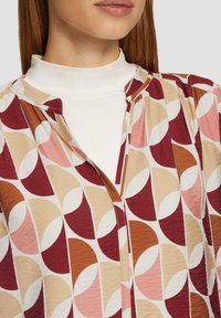 Overhemd met knoopsluiting en een geometrisch patroon in rood, beige en roze tinten. Heeft een hoge kraag en plooidetails bij de halslijn.