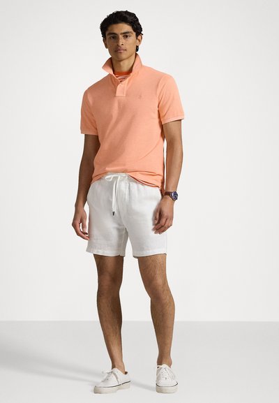 Polo arancione con colletto ripiegato, abbinato a pantaloni corti bianchi in lino e sneaker bianche. Il modello sta in piedi, mostrando i dettagli dell'outfit.