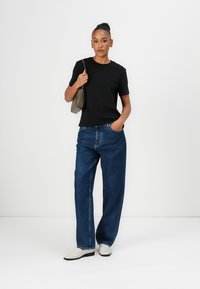 Svart bomullst skjorte, løstsittende blå denimjeans og hvite ankelstøvler. Modellen holder en liten grå håndveske, stående mot en nøytral bakgrunn.