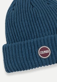 Pletená čiapka v tmavomodrej farbe, s rebrovanou textúrou a preloženým lemom. Má okrúhlu odznak značky Colmar v bielej farbe s vínovými akcentmi.