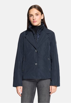 Vrouw die een donkerblauw dubbelbreasted jasje met revers draagt over een zwarte coltrui en grijze spijkerbroek, staand tegen een witte achtergrond.